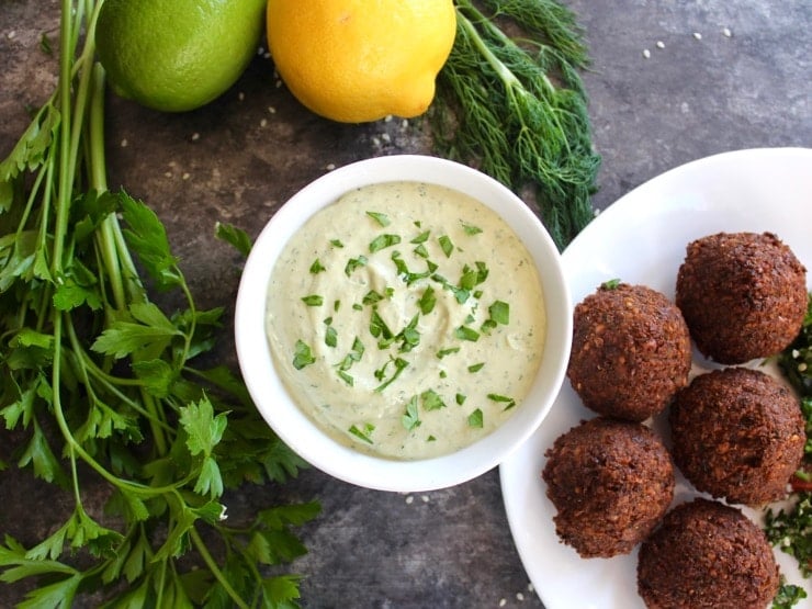 White bowl of herb tahini sauce topped with chopped fresh parsley alongside a falafel plate, fresh parsley, fresh dill, lemon and lime on the side.