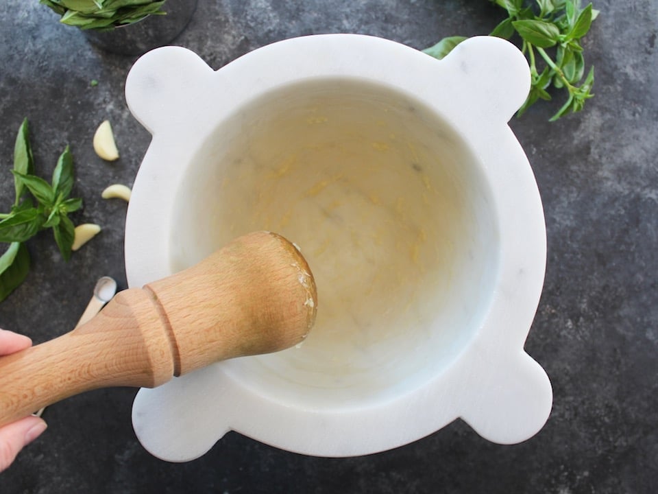 Close up horizontal shot of hand holding a wooden pestle over a white marble mortar with mashed garlic clove.