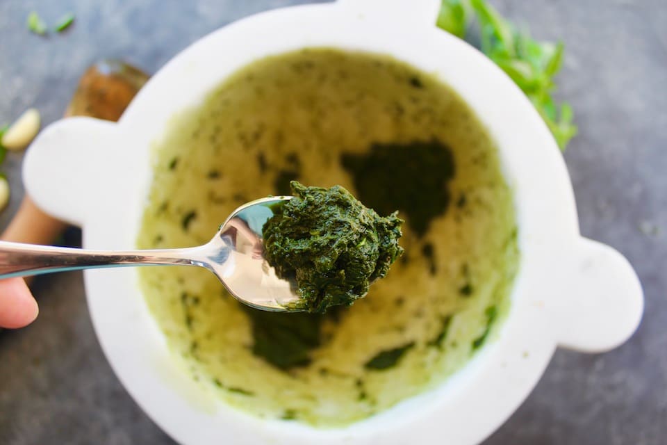 Close up of a spoonful of ground basil leaves held over a white marble mortar.