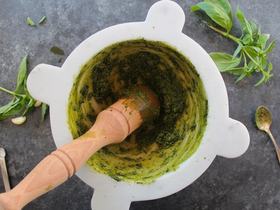 Close up of a wooden pestle grinding basil leaves in a white marble mortar.