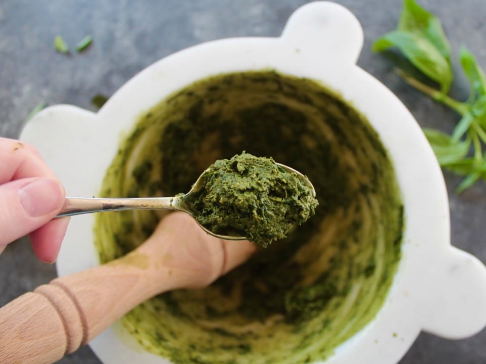 Close up of a spoonful of ground basil leaves held over a white marble mortar.