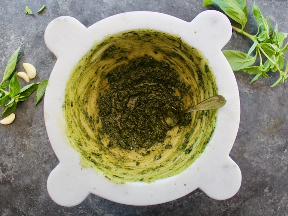 Stirring olive oil into the basil pesto in a marble mortar on countertop, basil herbs and garlic beside it.