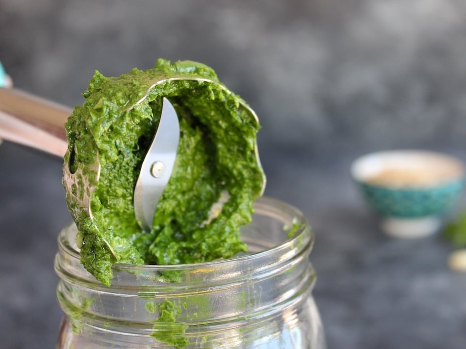 Close up shot of immersion blender blades covered in fresh basil pesto, held over the opening of a glass mason jar.