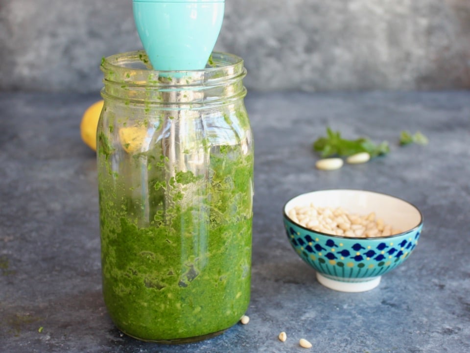 Horizontal shot of an immersion blender inside of a glass mason jar filled with fresh basil pesto. A decorative bowl sits in the background to the right.