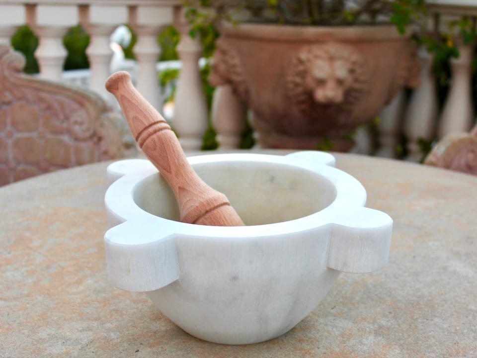 Traditional Italian marble mortar and wooden pestle on a stone table outside, chair and planter with lion\'s head in background, balustrades in background.