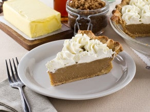 Slice of Butterscotch Pie topped with whipped cream and served on a white plate with a butter, fork, and glass of brown sugar on the side.