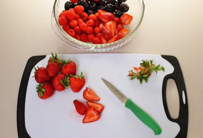 Overhead shot of sliced and hulled strawberries and a green paring knife sitting on top of a cutting board next to a bowl of berries.
