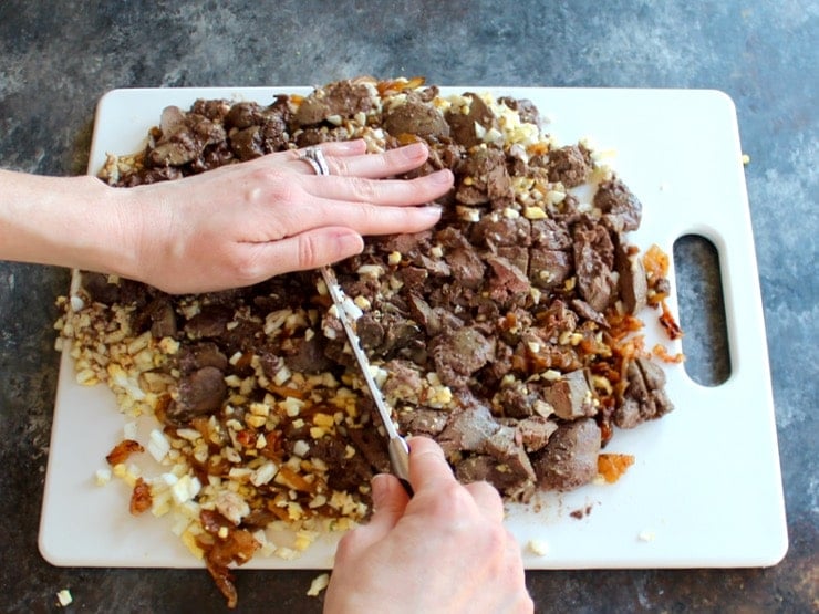 Hand chopping chicken livers, egg, and gribenes on a cutting board with a large chef's knife.