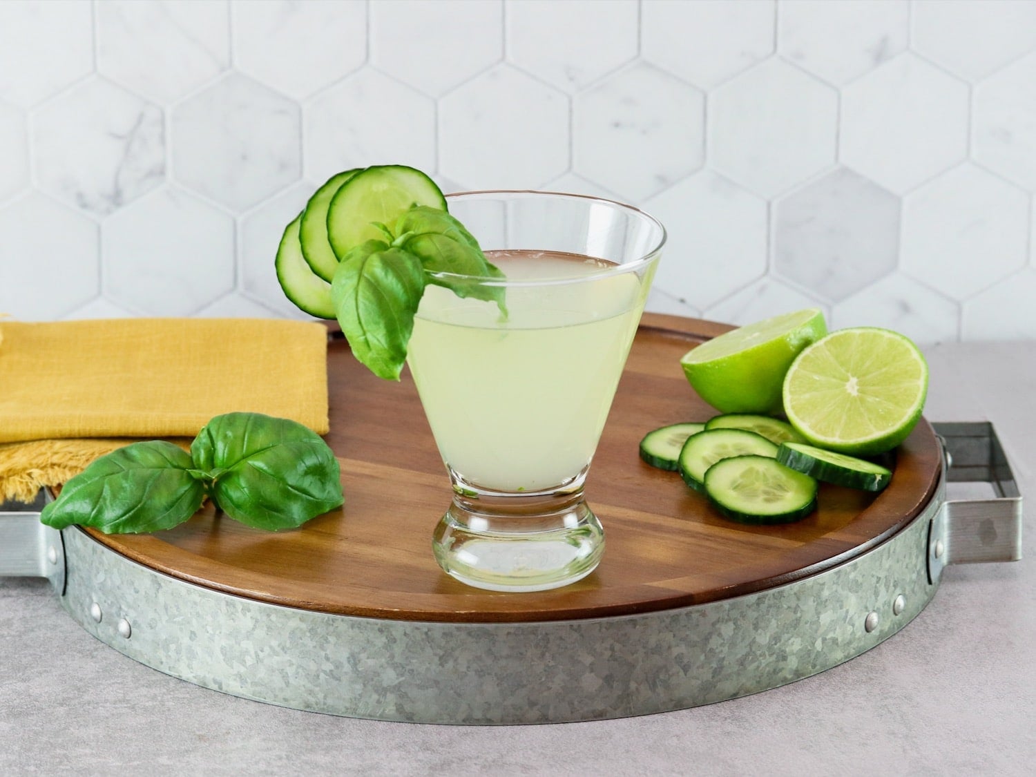 A short cocktail glass filled with a pale green drink and topped with cucumber slices and fresh mint.