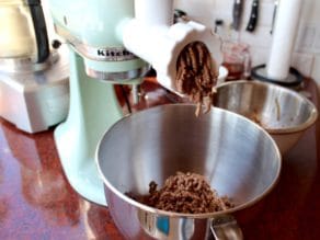 Mixer grinding chicken livers with meat grinder attachment into stainless steel bowl, another bowl and kitchen implements in background.