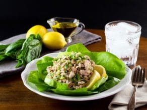 Healthy tuna salad on bed of green butter lettuce with lemon slices, on white plate with fork and cloth napkin, glass of ice water, lemons basil and olive oil in background.