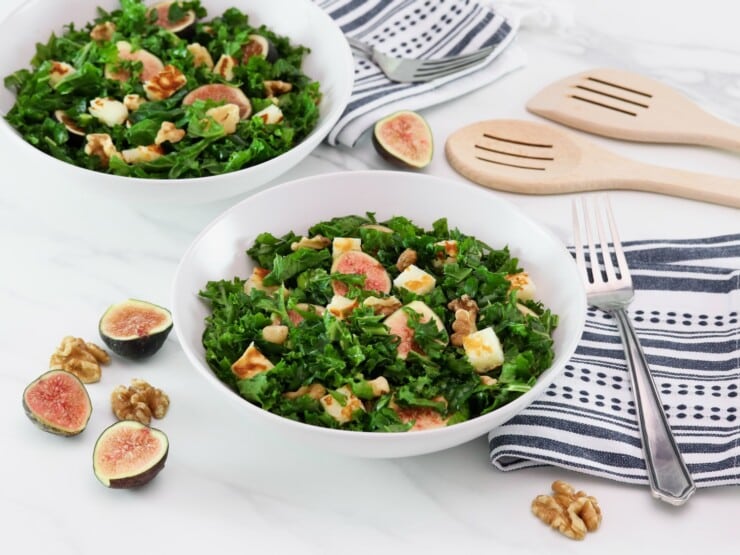 Overhead tilted horizontal shot of two white bowls filled with fig salad - fresh fig halves tossed with dark green curly kale, fried halloumi cheese, and walnuts. Linen napkins and fork, two wooden spoons for serving decorate the white marble countertop, alongside fresh fig halves scattered beside the bowls.