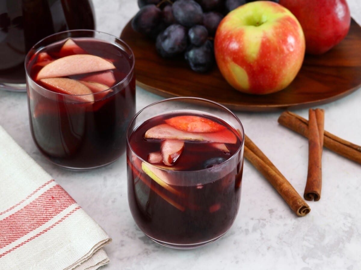 Tilted overhead shot of two glasses of red wine Thanksgiving sangria with fruit slices, three cinnamon sticks on the counter, a tray of fruit - apples and grapes, and a pitcher of sangria in the background. Linen towel on the side.