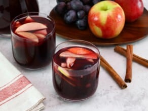 Tilted overhead shot of two glasses of red wine Thanksgiving sangria with fruit slices, three cinnamon sticks on the counter, a tray of fruit - apples and grapes, and a pitcher of sangria in the background. Linen towel on the side.
