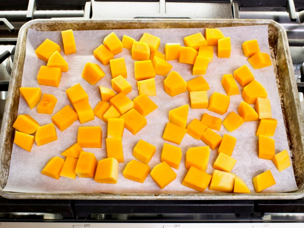 Overhead shot - tray of butternut squash cubes on a parchment-lined baking sheet, resting on a stovetop.