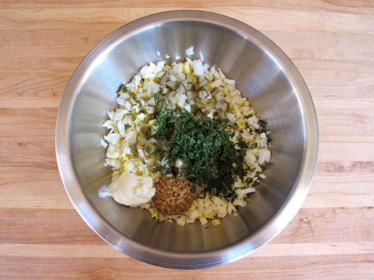 A metal bowl on a wooden countertop containing chopped eggs, dill, mustard, and mayonnaise. The ingredients are ready to be mixed for a salad.