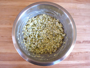 A metal bowl on a wooden countertop containing mixed ingredients of chopped eggs, dill, mustard, and mayonnaise.