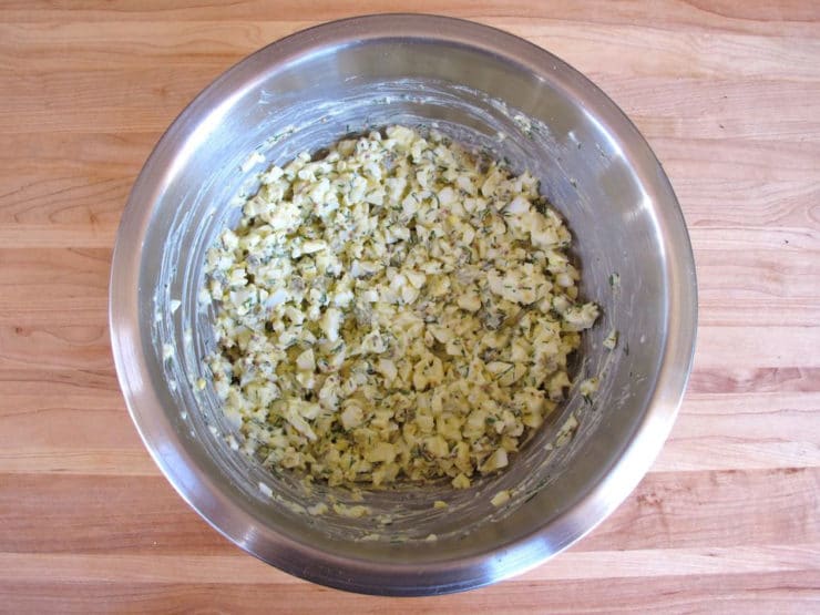 A metal bowl on a wooden countertop containing mixed ingredients of chopped eggs, dill, mustard, and mayonnaise.