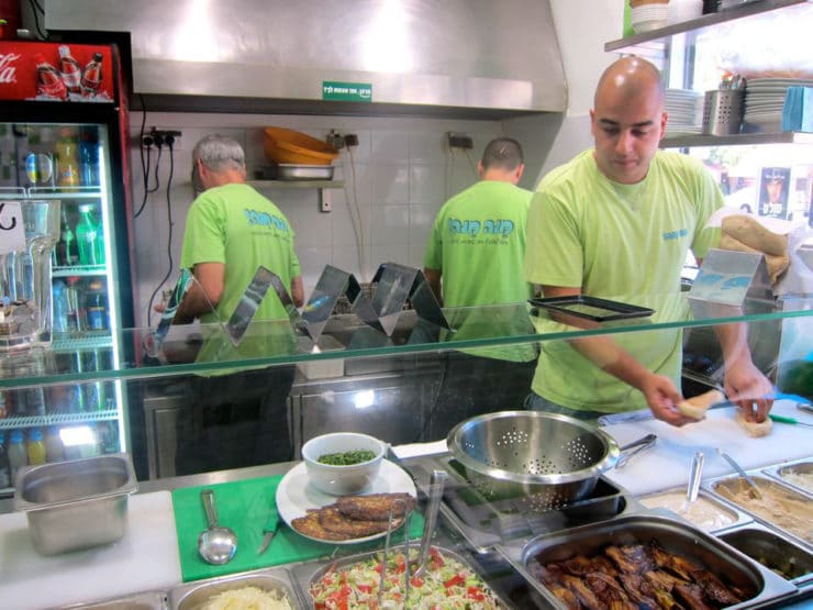 A man is making a pita sandwich at counter with salad fixins. Two workers are in the kitchen behind him.