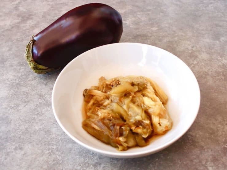 Roasted eggplant pulp in bowl with whole eggplant in background on grey background. 