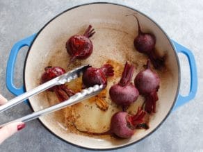 Flipping half-cooked beets in enameled casserole dish with stainless tongs on a concrete background.
