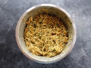 Shredded potato pancake batter mixed with tahini, herbs and spices in a large mixing bowl on concrete background.