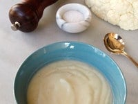 Vertical image of Cauliflower Soup in a blue bowl with a golden spoon, small bowl of salt, cauliflower, and pepper shaker.