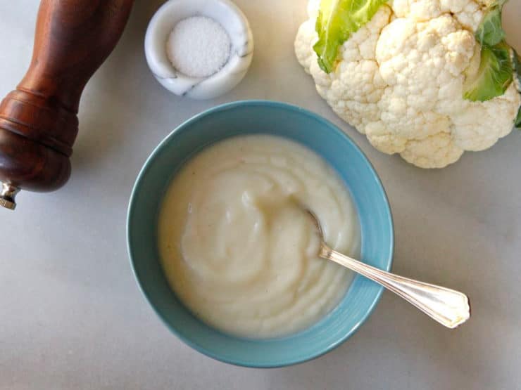 A bowl of creamy cauliflower soup garnished with a spoon accompanied with cauliflower, salt, and pepper on the side