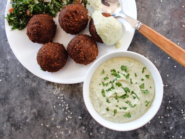 White bowl of herb tahini sauce topped with chopped fresh parsley alongside a falafel plate. One piece of falafel topped with tahini sauce, spoon beside it. Tabouli salad in background.