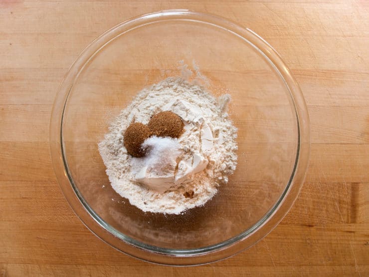A glass bowl on a wooden surface contains a mixture of flour, brown sugar, and salt, ready for baking.