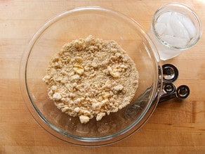 Glass bowl with crumbly dough on a wooden surface, next to measuring spoons and a glass of ice water.