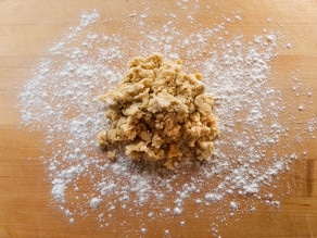 A mound of raw dough sits on a wooden surface, surrounded by scattered white flour.