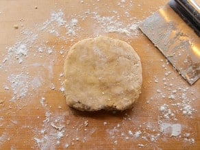 A ball of dough rests on a floured wooden surface next to a metal dough scraper.