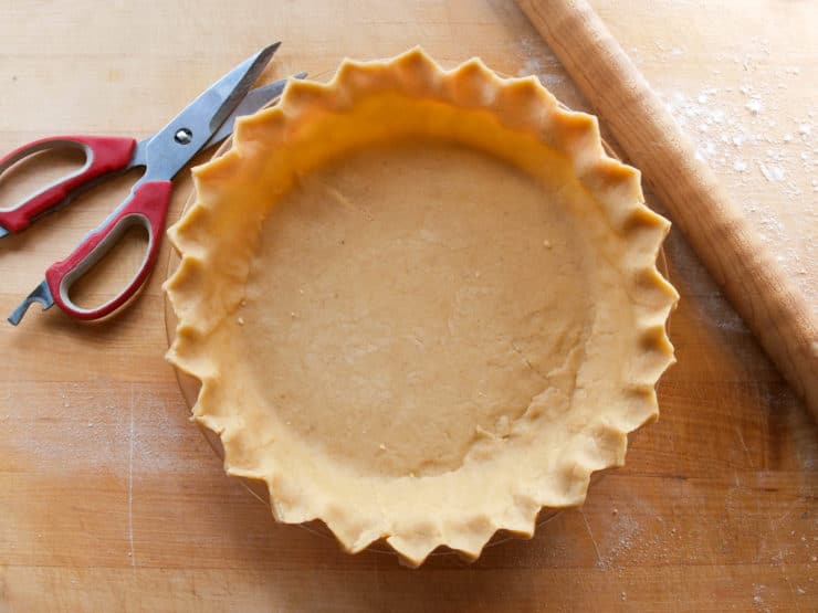 A fluted pie crust in a pan is on a wooden surface, next to red-handled scissors and a dusted rolling pin.