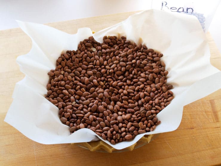 A pie crust lined with parchment paper is filled with dry beans, resting on a wooden surface.