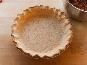 Golden-brown pie crust in a glass dish on a wooden surface, next to a bowl of dried beans.