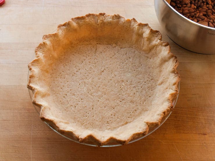 Golden-brown pie crust in a glass dish on a wooden surface, next to a bowl of dried beans.