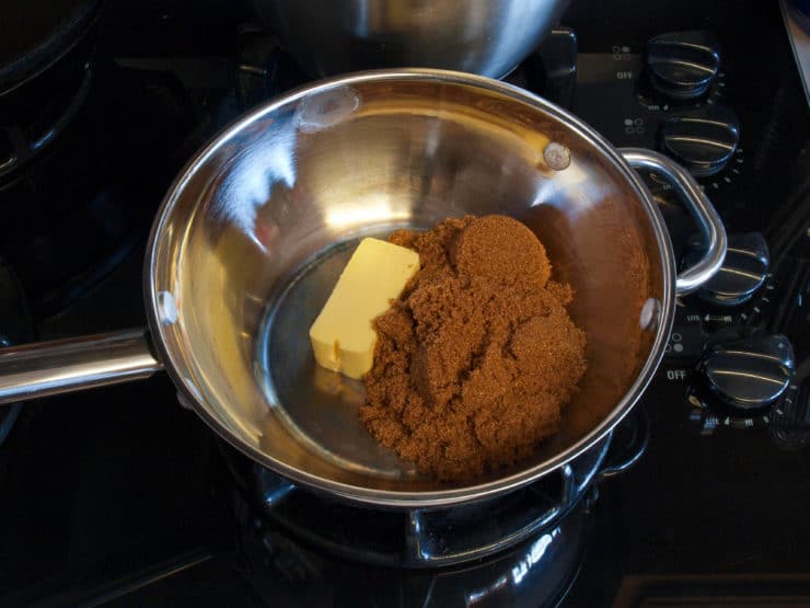 A stainless steel pan on a stovetop contains a block of butter and a mound of brown sugar, ready for melting.