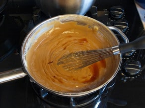 A saucepan on a stove contains smooth, creamy caramel sauce with vanilla being whisked