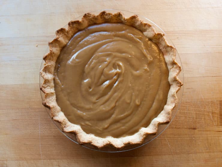 Butterscotch Pie with a golden, fluted crust sits on a wooden surface. The pie's smooth filling is swirled, conveying a warm, homemade feel.