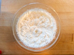 A glass bowl filled with whipped cream sits on a wooden table. The cream is thick and fluffy, forming soft peaks, suggesting it's been freshly whipped.