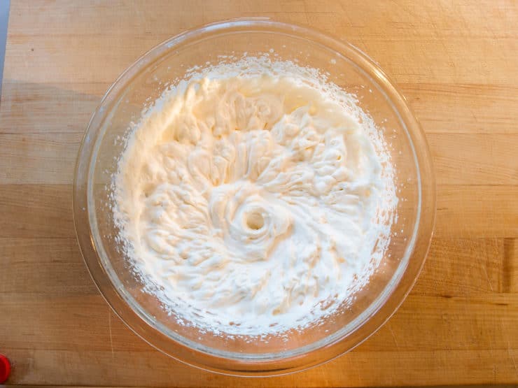 A glass bowl filled with whipped cream sits on a wooden table. The cream is thick and fluffy, forming soft peaks, suggesting it's been freshly whipped.