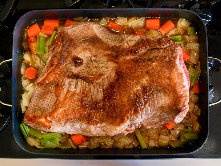 Browned brisket on top of onions, carrots and celery chopped in roasting pan on stovetop.
