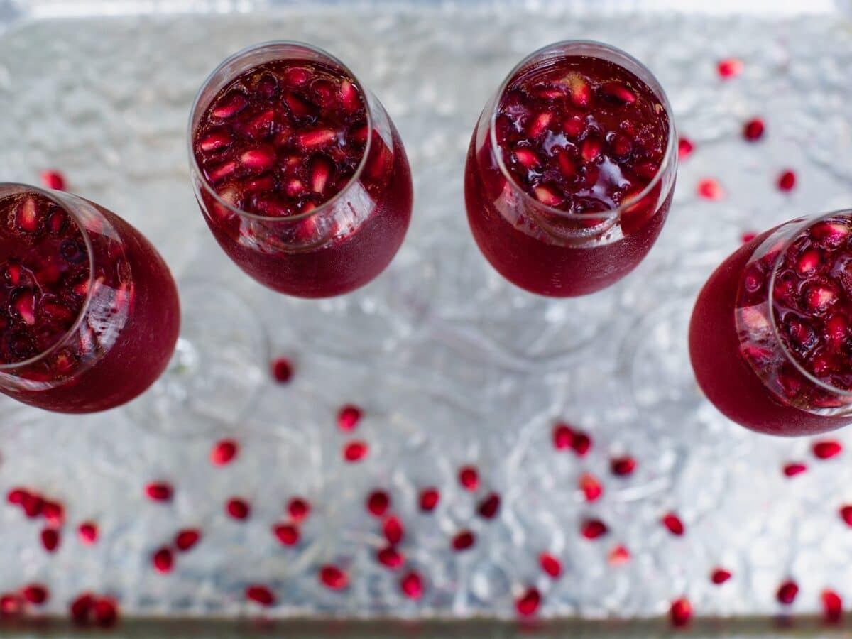 Overhead horizontal shot of four champagne flutes filled with a bright red pomegranate cocktail. Pomegranate arils are floating in the cocktails and scattered around the table.