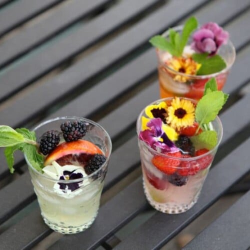 Three shot glasses filled with drinks and topped with berries, mint, and beautiful small flowers