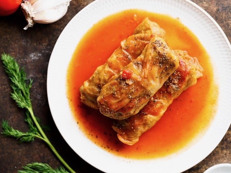 Three stuffed cabbage leaves on a plate with sauce, fresh dill and garlic on the side.