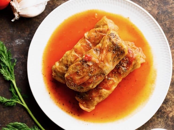 Three stuffed cabbage leaves - holishkes on a plate with sauce, fresh dill and garlic on the side.