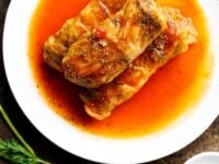 Stuffed Cabbage Leaves Pinterest Pin - a plate of three stuffed cabbage leaves on a plate with tomato sauce, sprinkled with cracked black pepper. Fresh dill and garlic on the countertop beside the plate.