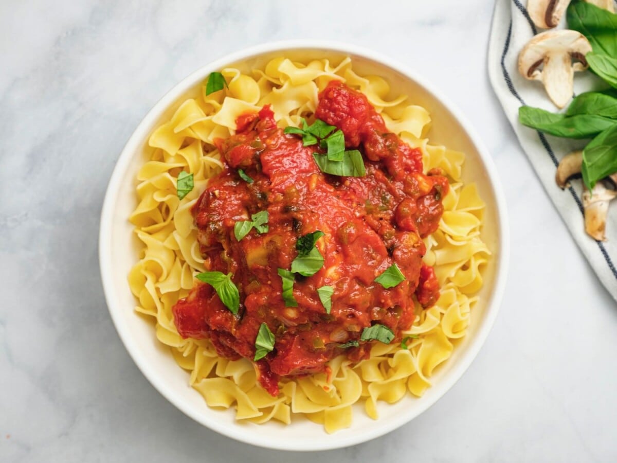 Overhead horizontal shot of a bowl egg noodles topped with chicken cacciatore in tomato sauce and garnished with parsley, on a marble countertop.
