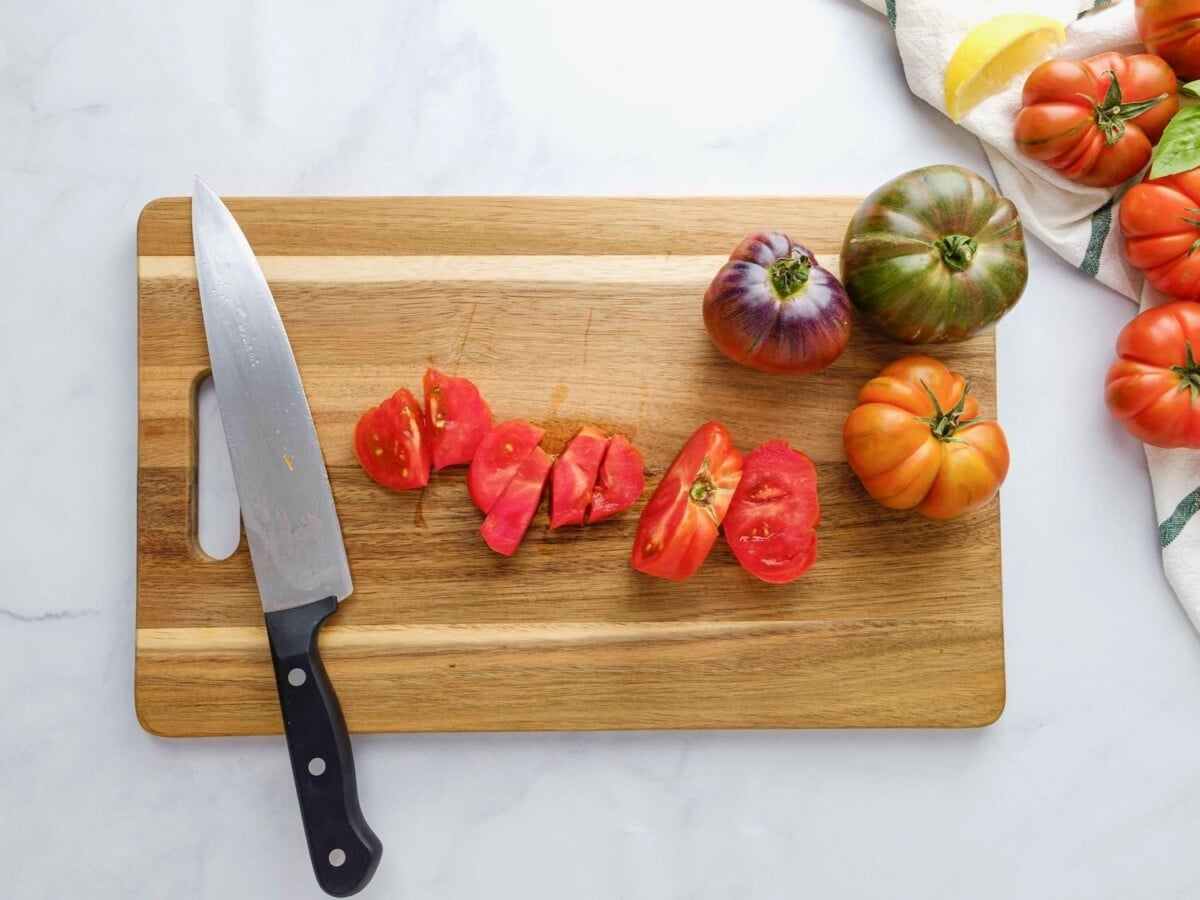 Sliced and chopped heirloom tomatoes with three whole heirloom tomatoes on a cutting board, with a chef's knife resting on the board. Fresh heirloom tomatoes, cloth towel, and lemon slices off to the side.